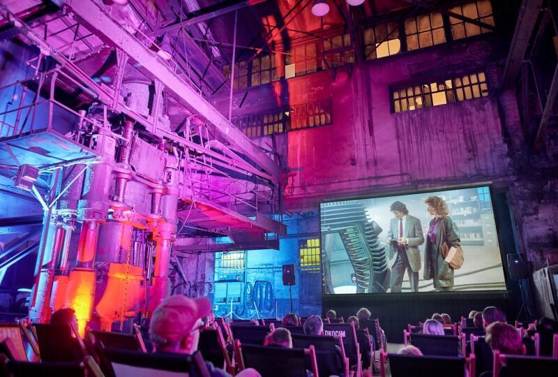 W środku w głębi ekran, na którym wyświetlają się kolorowe postacie, przed ekranem ludzie siedzą na krzesłach, fotelach kinowych 