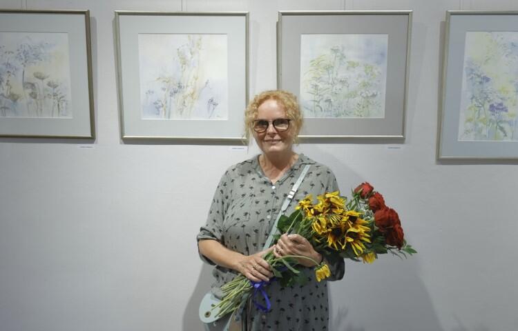 Starsza kobieta w okularach, blondynka trzyma w ręku bukiet kwiatów, w tle obrazy