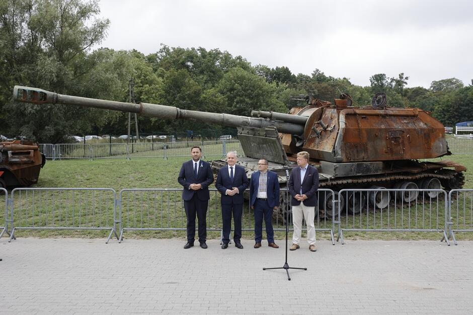 Czterech mężczyzn w róznym wieku w garnitura stoi przed mikrofonami, za nimi zniszczony czołg