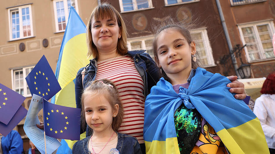Z prawej stoi dziewczynka w żółto niebieskiej fladze na ramionach, w środku młoda kobieta, z lewej strony mała dziewczynka 