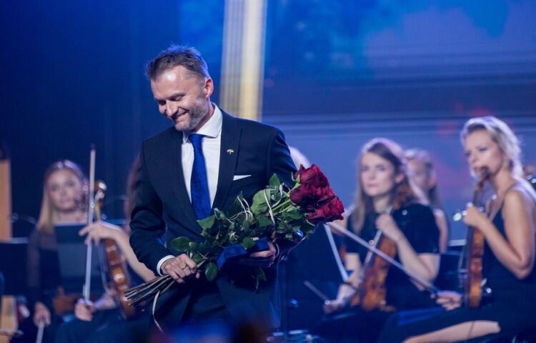 Młody uśmiechnięty mężczyzna z bukietem czerwonych róż na scenie, za jego plecami skrzypaczki z orkiestry