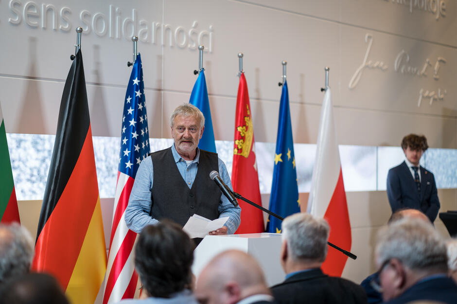 na zdjęciu starszy mężczyzna w koszuli i ciemnej kamizelce, stoi za jasnym pulpitem, przemawia, przed nim siedzi grupa osób, widać ich głowy, za mężczyzną widać kilka flag, między innymi polską, unii europejskiej i gdańska