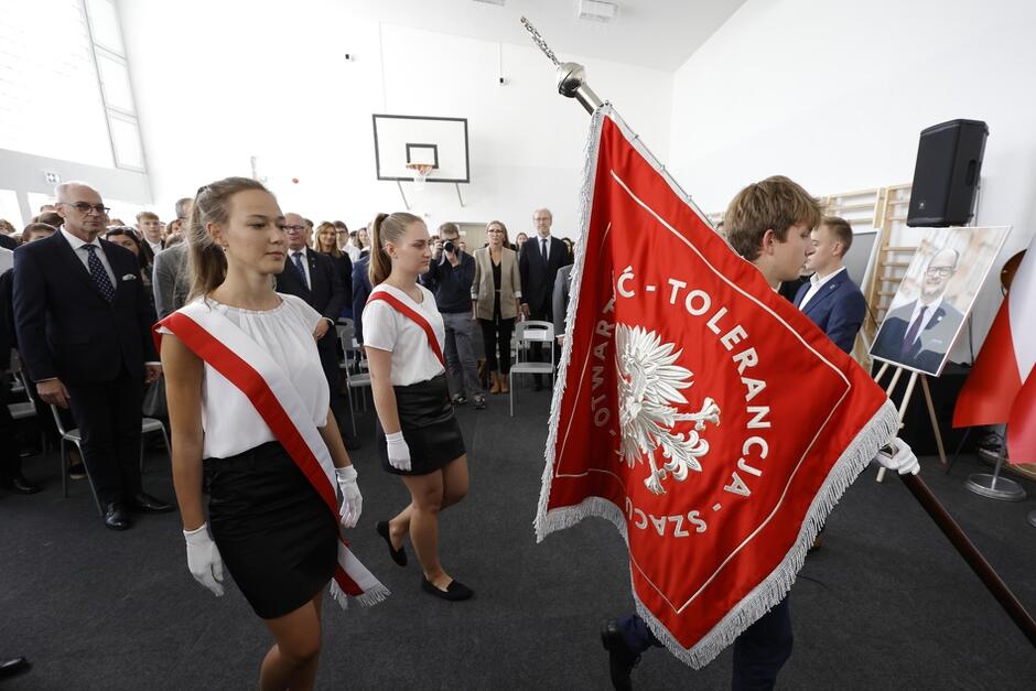 sala gimnastyczna, w centrum kadru poczet sztandarowy szkoły, dwie dziewczyny przepasane flagą polski idą za chłopakiem niosącym czerwony sztandar z wyhaftowanym orłem w koronie i słowami szacunek, otwartość, tolerancja