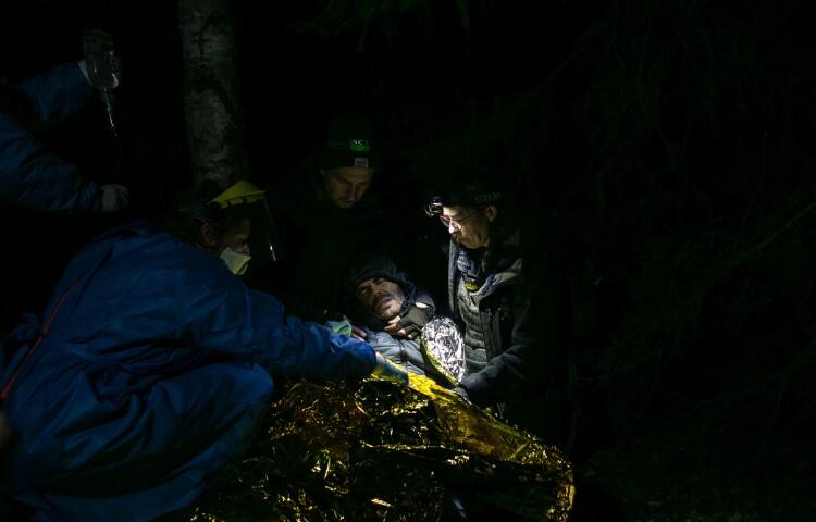 Zdjęcie wykonane w nocy. Na ziemi leży człowiek przykryty folią ratunkową, nad nim pochylają się lekarze i sanitariusze, z ciemności wyłania się cierpiąca twarz chorego 