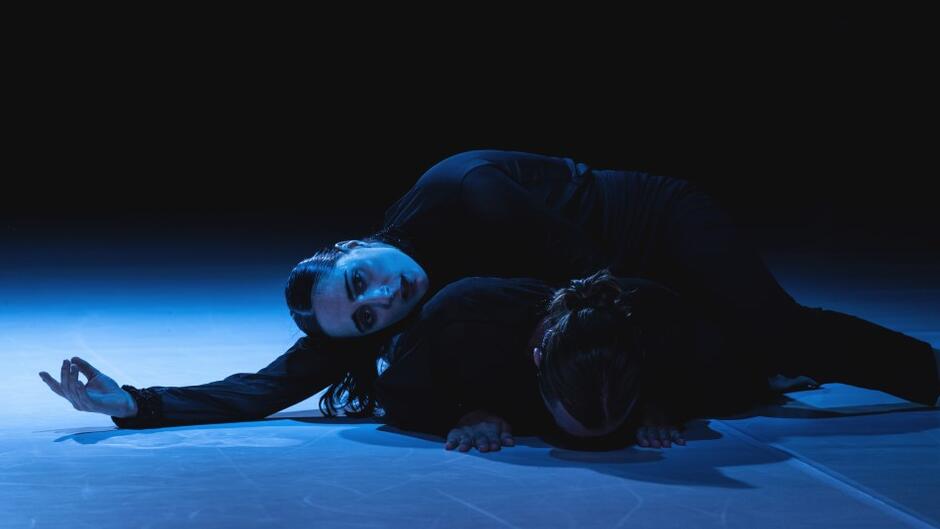 dwie tancerki w ciemnej scenerii, dziewczyna ma wysunięte ramię, leży na ziemi, patrzy w obiektyw