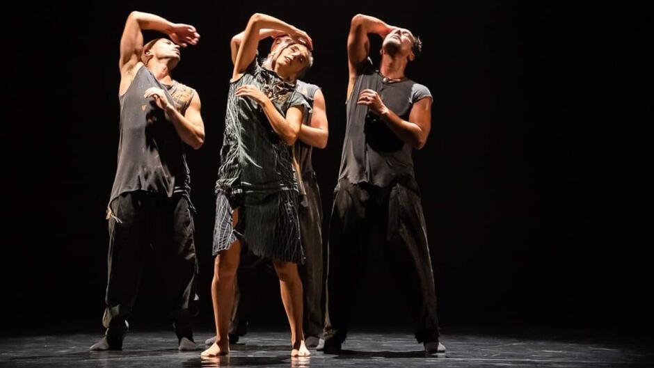 czterej tancerze, jedna kobieta w szarych poszarpanych strojach stoją na scenie blisko siebie w tej samej pozycji. jedna ręka sięga za głowę, druga jest blisko ciała