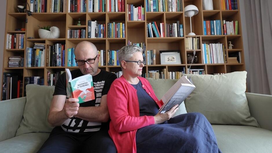 mężczyzna i kobieta w okularach, lekko siwiejący, obcięci krótko po męsku. siedzą na kanapie opierając się o siebie, mają w ręku książki, są zaczytani 