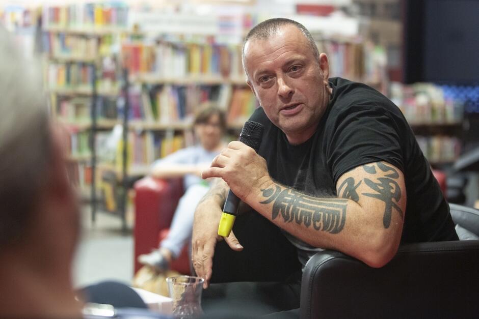 mężczyzna w czarnym T-shirtcie z krótkimi rękawami, ma wytatuowane ramiona, trzyma w ręku mikrofon, w tle półki z książkami