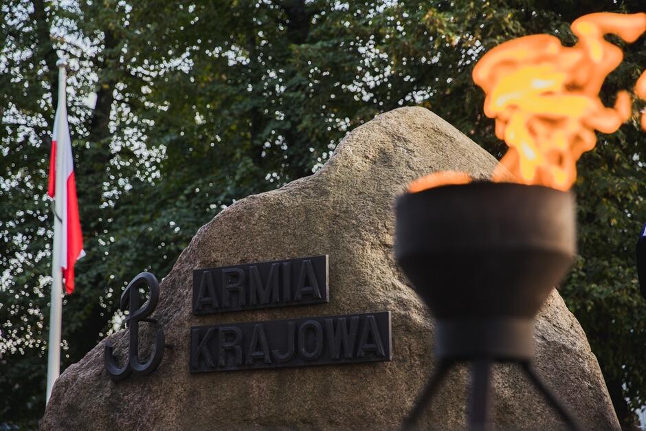 Widok pomnika w postaci granitowego głazu narzutowego z wykonanym w metalu znakiem Polska Walcząca i napisem Armia Krajowa. Na prawo od głazu znajduje się duży płonący znicz 