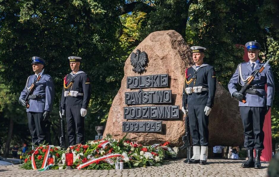 Pomnik Polskiego Państwa Podziemnego w Gdańsku, koło niego stojący żołnierze Marynarki Wojennej, przed pomniniem leżą wieńce wykonane  z kwiatów