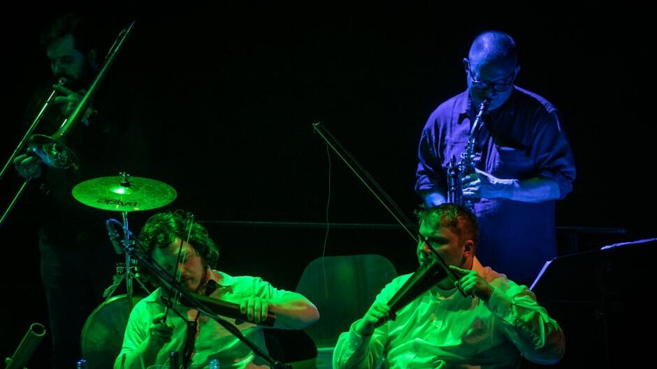 Zdjęcie w kolorach świateł, muzyk stoi i gra na saksofonie, przed nim dwie osoby w białych koszulach siedzą za stołem z instrumentami perkusyjnymi w rękach