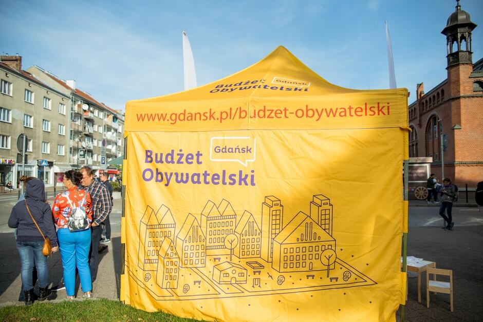duży żółty namiot z napisem: Budżet Obywatelski ustawiony na ulicy w centrum miasta, obok niego stoi grupa czterech osób