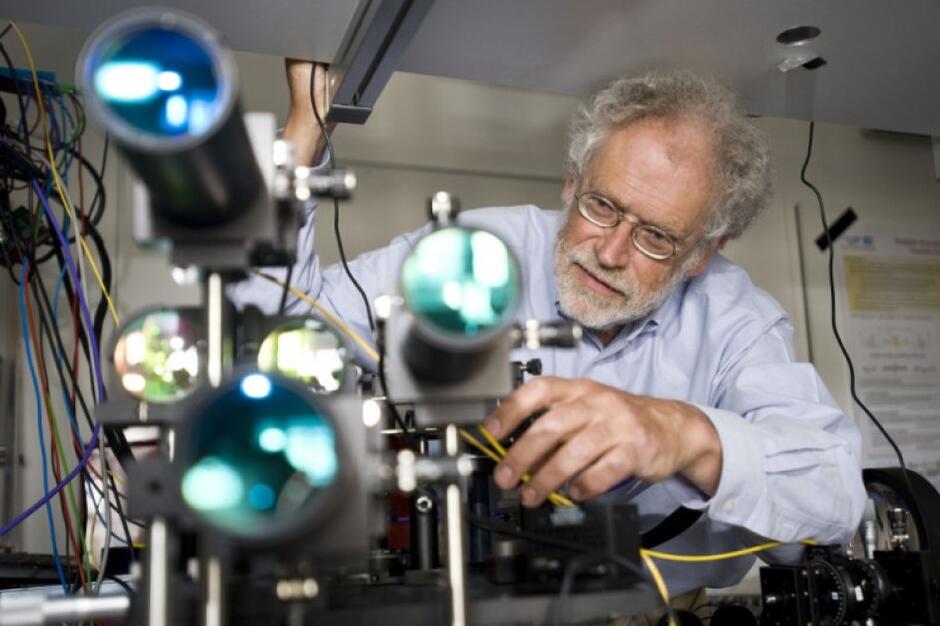 Mężczyzna w średnim wieku z siwymi włosami w centrum obok niego próbówki, aparatura naukowa