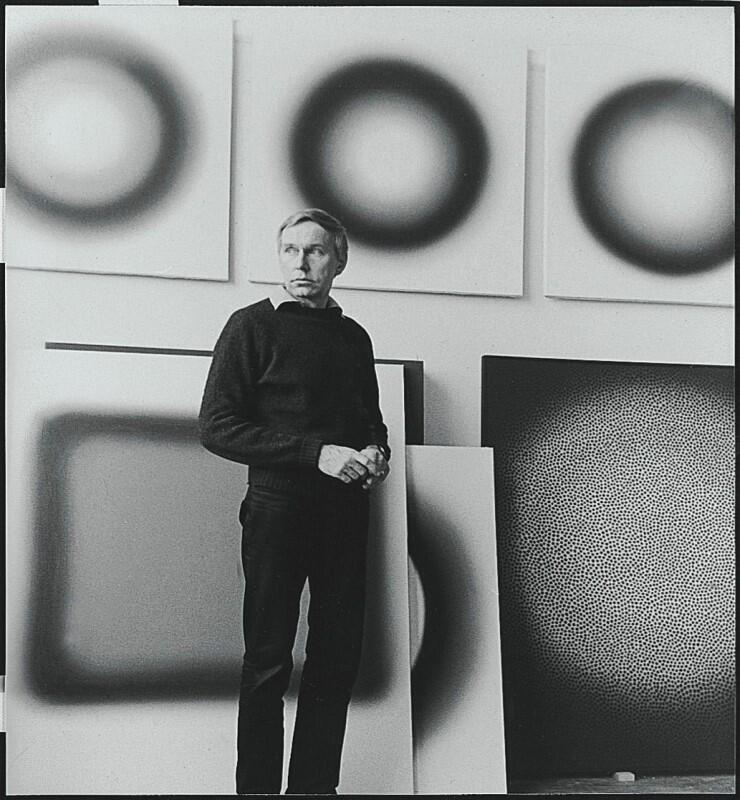 czarno białe zdjęcie, szczupły mężczyzna stoi na tle abstrakcyjnych obrazów