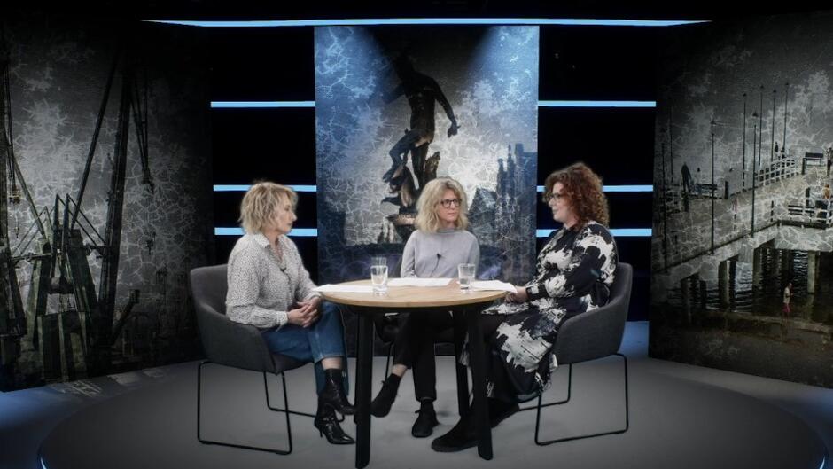 trzy kobiety siedzą w studiu, przy okrągłym stole. W tle wizerunki miasta