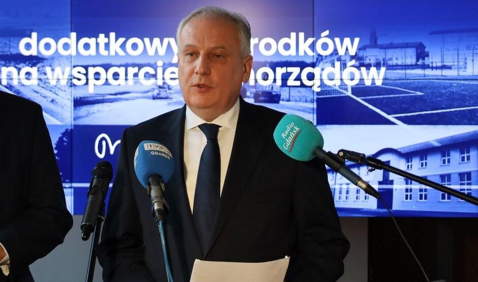 Siwy mężczyzna w garniturze pod krawatem. Przed nim mikrofony