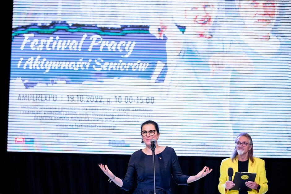 Młoda kobieta w sukience i okularach stoi przed mikrofonem, za nią na ekranie wyswietla się napis Festiwal Pracy i Aktywności Seniorów. Z prawej strony w głębi stoi mężczyna w średnim wieku z długimi włosami