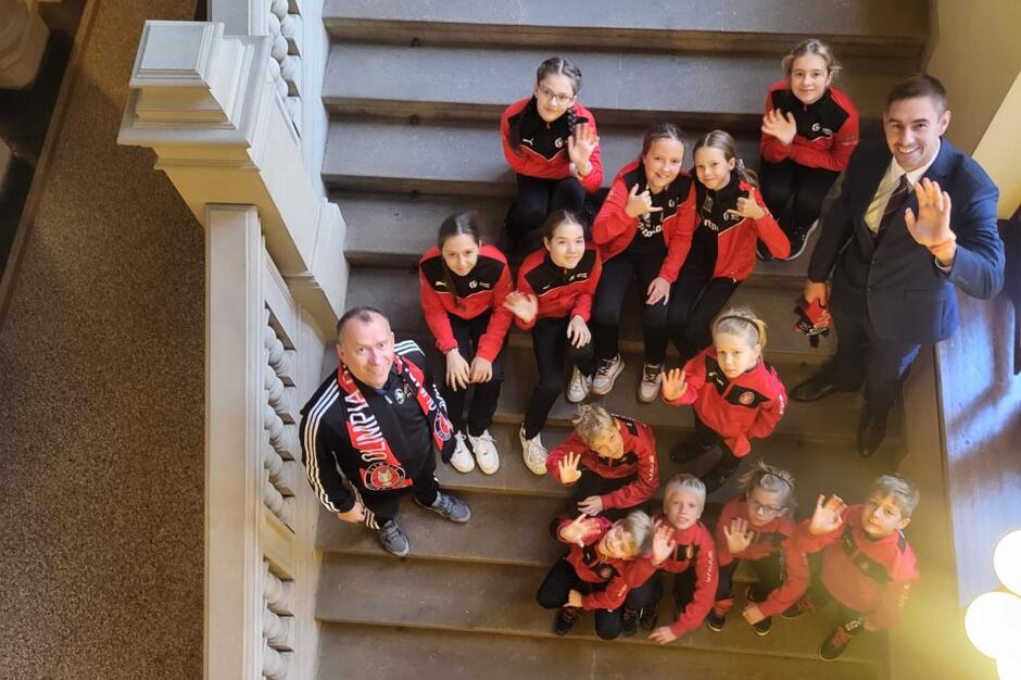 Kilkanaście dziewcząt i chłopców w wieku szkolnym - stoją na wewnętrznych schodach w gmachu Rady Miasta Gdańska. Towarzyszy im dwóch opiekunów. Wszyscy patrzą w górę w stronę fotografa i machają rękami w geście pozdrowienia. Zdjęcie zrobione z wyższego piętra 