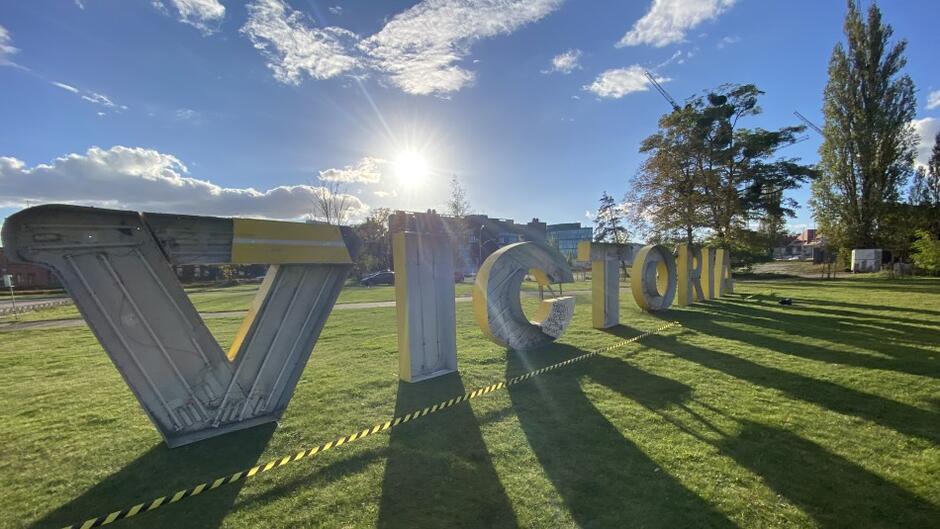 "Victoria" - napis stoi w słońcu i rzuca cień na trawniku na placu Porozumień Gdańskich