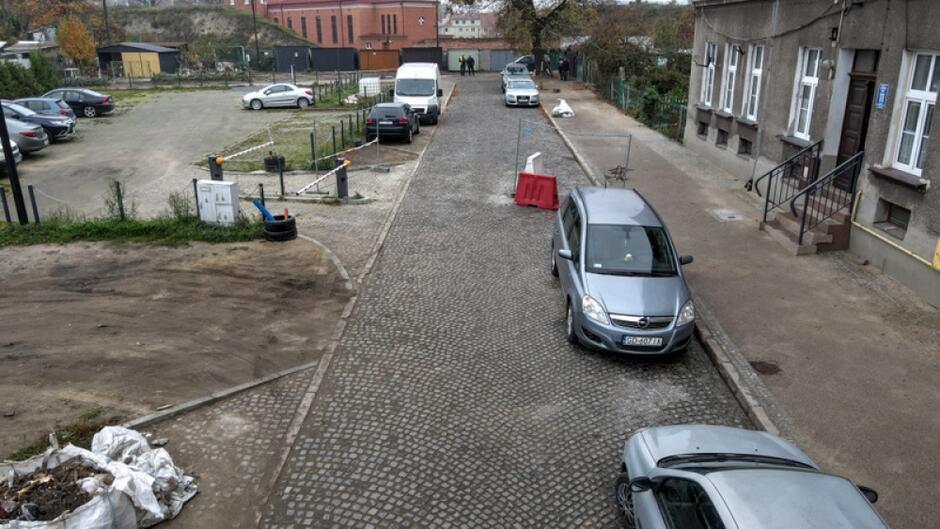 na zdjęciu z drona fragment brukowanej ulicy, po prawej stoją zaparkowane samochody osobowe, po prawej fragment szarej kamienicy