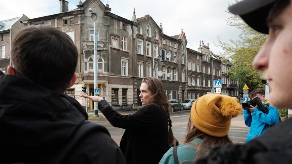 grupa ludzi stoi na ulicy, jedna z kobiet wskazuje na fasadę historycznego budynku