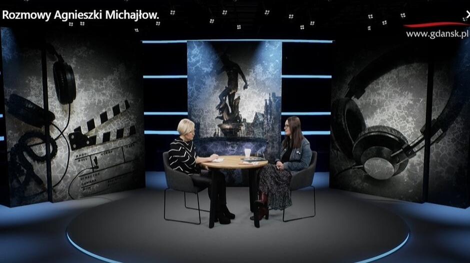 Z prawej strony młoda kobieta z długimi włosami siedzi na krześle przy okrągłym stole, z lewej kobieta w średnim wieku w okularach siedzi na krześle. Z tyłu, za nimi, na ekranie napisy Jestem z Gdańska 