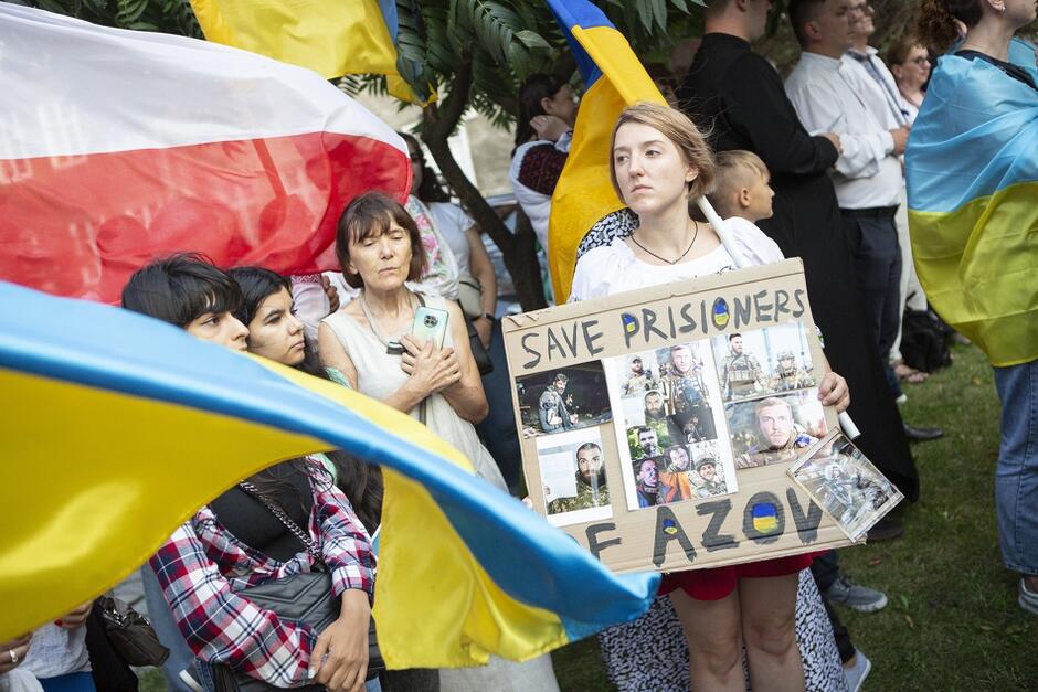Grupa osób podczas obchodów święta niepodległości Ukrainy. Stoją, trzymając niebiesko-żółte flagi. Wśród nich jest też duża flaga biało-czerwona. Jedna z kobiet trzyma tablicę wzywająca do uwolnienia obrońców Azovstalu w Mariupolu