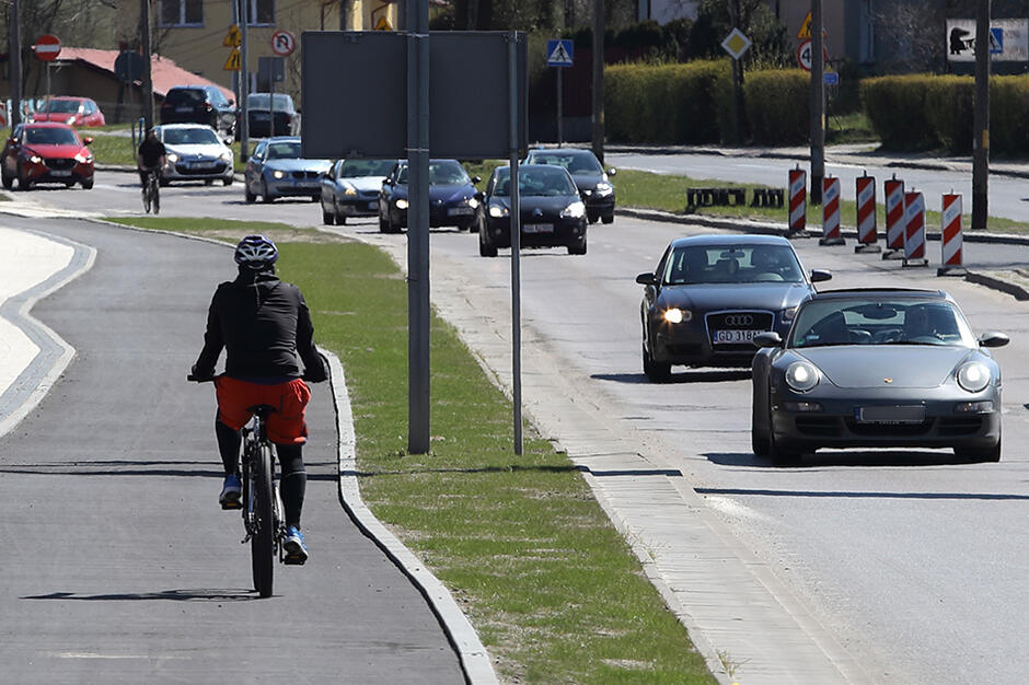 Z prawej strony ulica i jadace po nie samochody, obok ścieżka rowerowa, po niej jedzie rowerzysta  
