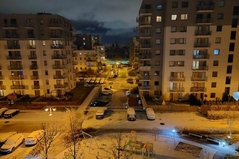 Zdjęcie zrobione z wysokiego piętra bloku mieszkalnego. Widać ulice i trawniki oraz samochody na parkingach - wszystko pokryte warstwą śniegu. Naprzeciwko osoby fotografującej są dwa inne bliki mieszkalne