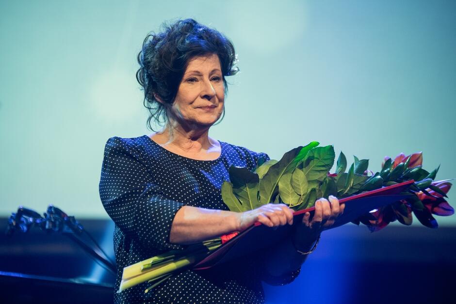 Dorota Kolak, kobieta ubrana w ciemną sukienkę, stoi na scenie, w dłoniach ma dyplom i kwiaty