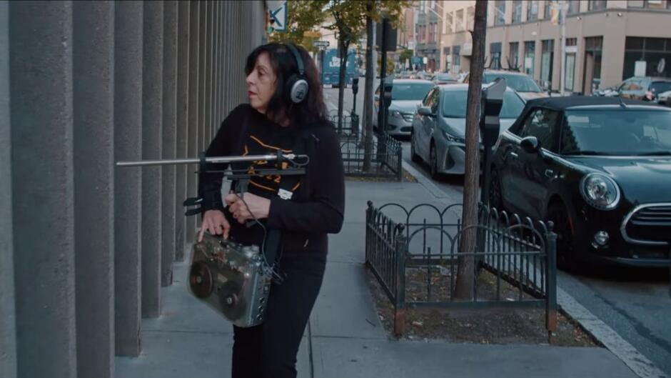 Kobieta z długimi ciemnymi włosami idzie ulicą miasta, ma przewieszony przez ramię magnetofon szpulowy, a w ręku mikrofon.