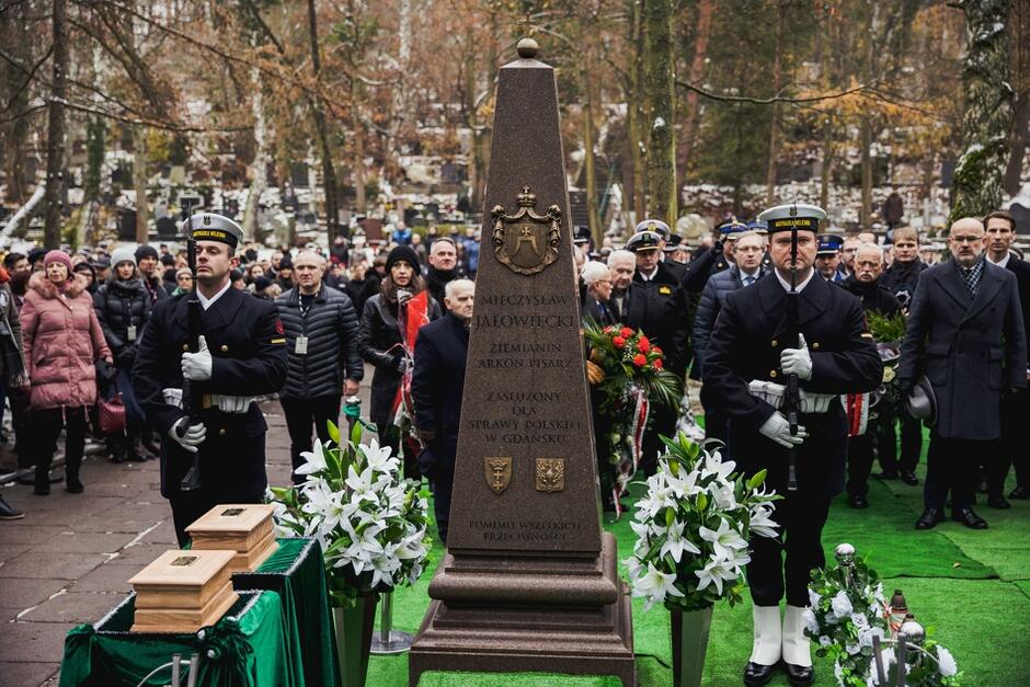 pogrzeb, dwóch żołnierzy marynarki wojennej stoi na baczność,  między nimi pomnik nagrobek, po obu stronach nagrobka  bukiety białych kwiatów w tle uczestnicy pogrzebu