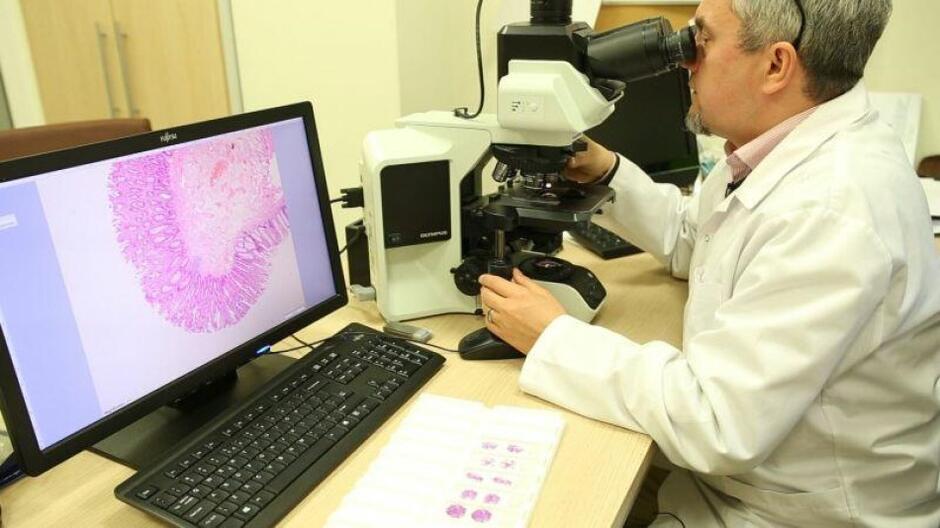 Z prawej strony mężczyzna w białym fartuchu siedzi i pochyla główę patrząc w mikrokop, na środku mikroskop, z lewej strony ekran, na którym widać obraz tkanek 