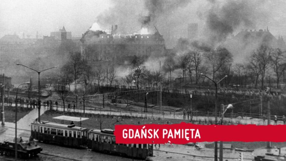 Na pierwszym planie tramwaj, za nim drzewa, w oddali kilkupiętrowy płonący budynek, czarno białe kolory, zdjęcie archiwalne. Z prawej strony napis Gdańsk pamięta 