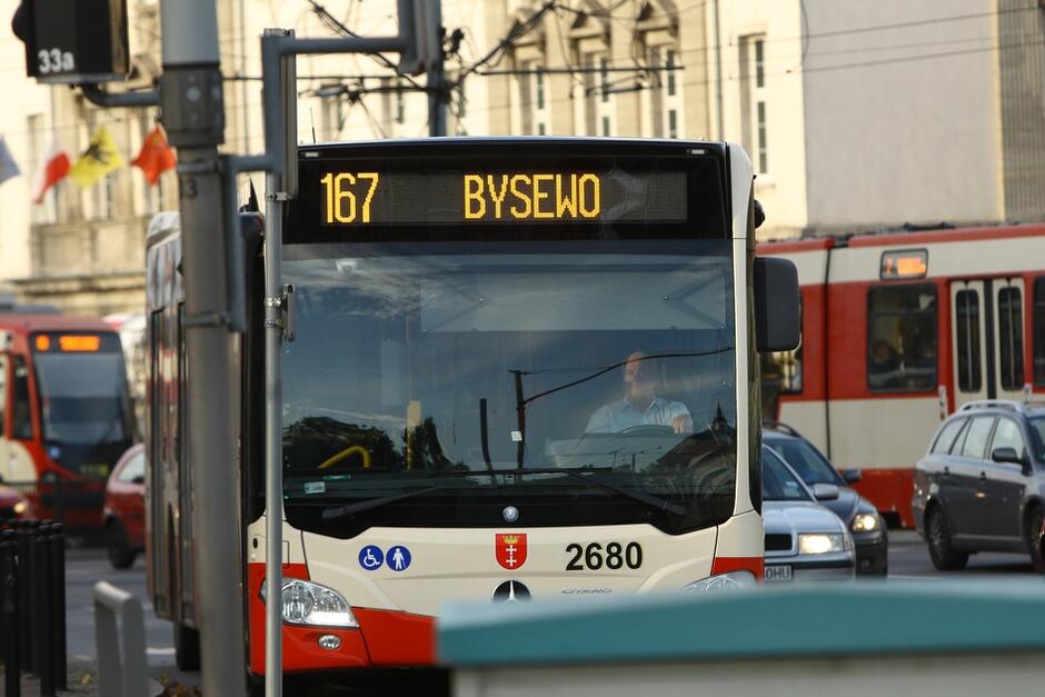 Zdjęcie miejskiej ulicy z dużą liczbą samochodów - wśród nich, na pierwszym planie jest autobus miejski, z napisem na wyświetlaczu pod dachem: 167 Bysewo. W tle widoczny miejski tramwaj