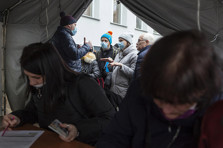 Namiot na świeżym powietrzu. W środku dwie kobiety siedzą przy stole, wypełniają dokumenty. Za nimi (przed wejściem do namiotu) stoi kilka innych osób 