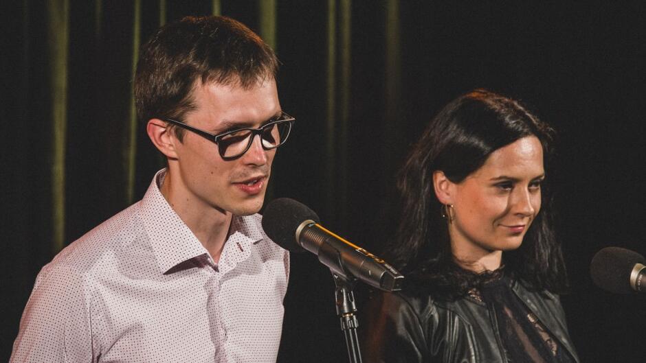 młody mężczyzna w okularach i kobieta - brunetka w ciemnym ubraniu - stoją na scenie, mówią do mikrofonu