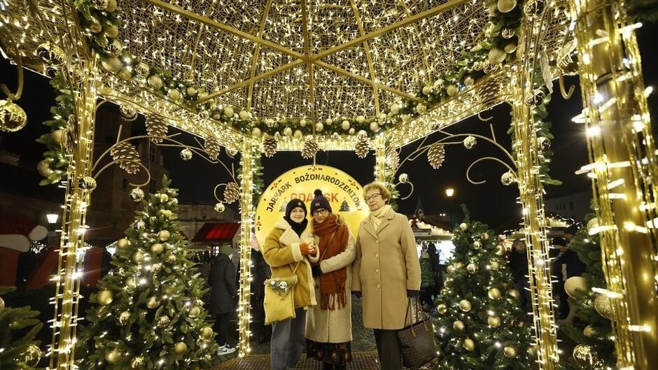 Trzy kobiety w ciepłych zimowych ubraniach pozują wewnątrz zbudowanej ze złotych światełek konstrukcji