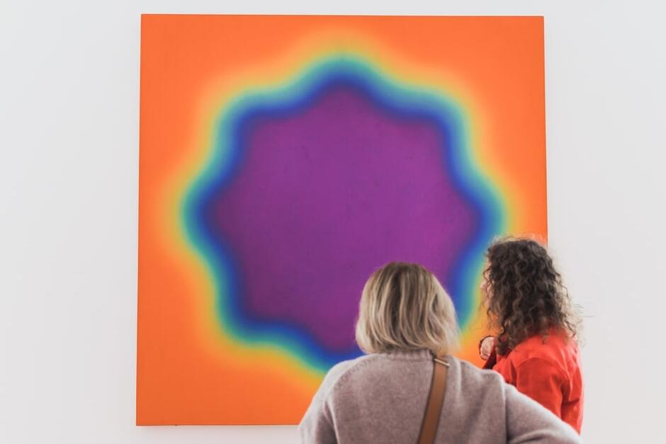 Przed abstrakcyjnym obrazem artysty - przestawiającym rozetę o przenikających się kolorach - stoją (do nas tyłem) dwie postaci kobiece 