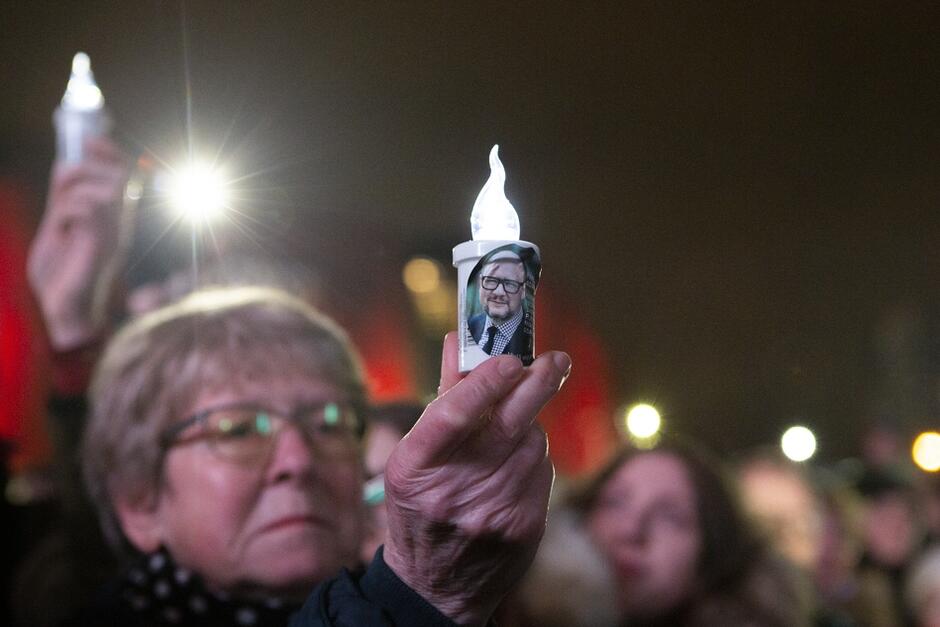 Tłum osób wieczorową porą. Na pierwszym planie starsza kobieta, która w dłoni trzyma świeczkę z naklejonym na nią portretowym zdjęciem Pawła Adamowicza 