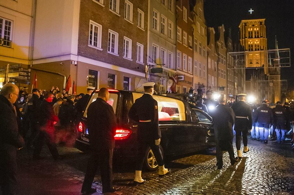 Kondukt wieczorem przemierza ulice Gdańska. Widzimy jadący samochód karawan, przez tylne szyby widać trumnę która znajduje się w tylnej części wnętrza auta - jest ona oświetlona 