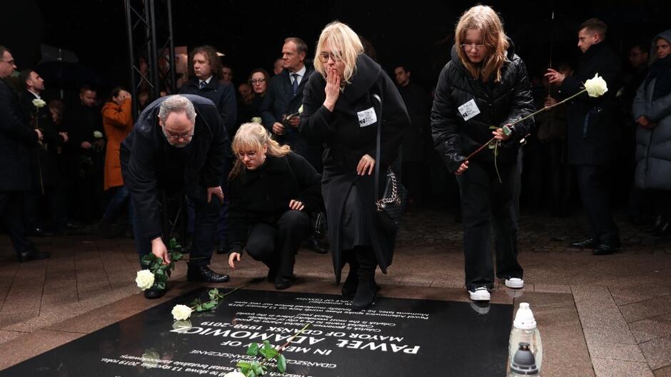 Trzy kobiety oraz mężczyzna - wszyscy ubrani na czarno - składają białe róże na płycie upamiętniającej miejsce zamachu na życie prezydenta Gdańska Pawła Adamowicza