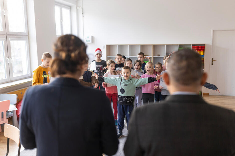 Kobieta i mężczyzna stoją tyłem w sali, przed nimi grupa dzieci
