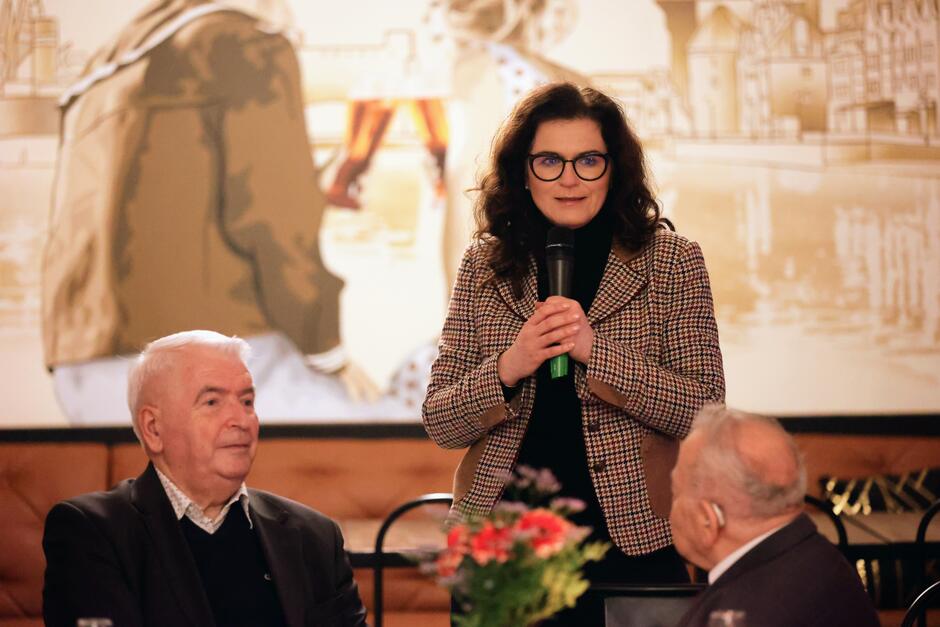na zdjęciu z przemawia do mikrofonu prezydent aleksandra dulkiewicz, przed nią siedzi na krzesłach dwóch starszych mężczyzn
