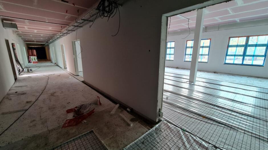 na zdjęciu fragment korytarza i sali lekcyjnej w powstającym budynku szkoły. widać że trwają prace budowlane, ściany są jasne, widać instalacje ułożone na podłodze, w tle widać kilka okien