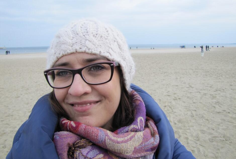 kobieta w czapce wełnianej i okularach uśmiecha się, w tle widać brzeg morza i plażę