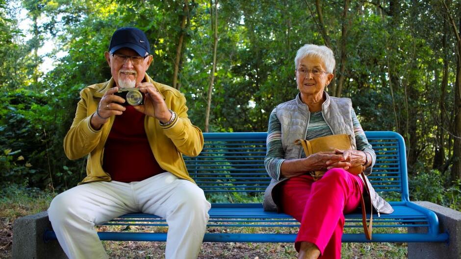 starszy mężczyzna i kobieta siedzą na ławce w parku w ciepły dzień, on trzyma przed sobą w dłoniach aparat fotograficzny, ona się uśmiecha
