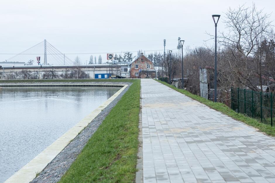 na zdjęciu nowy wał przeciwpowodziowy, widać chodnik, umocnienia od strony brzegu i fragment rzeki, w tle zabudowania i most wantowy