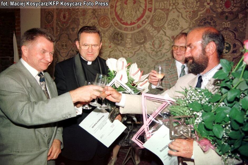 czterech mężczyzn, w tym jeden w sutannie, stoją w zabytkowym wnętrzu trzymając duży bukiet kwiatów