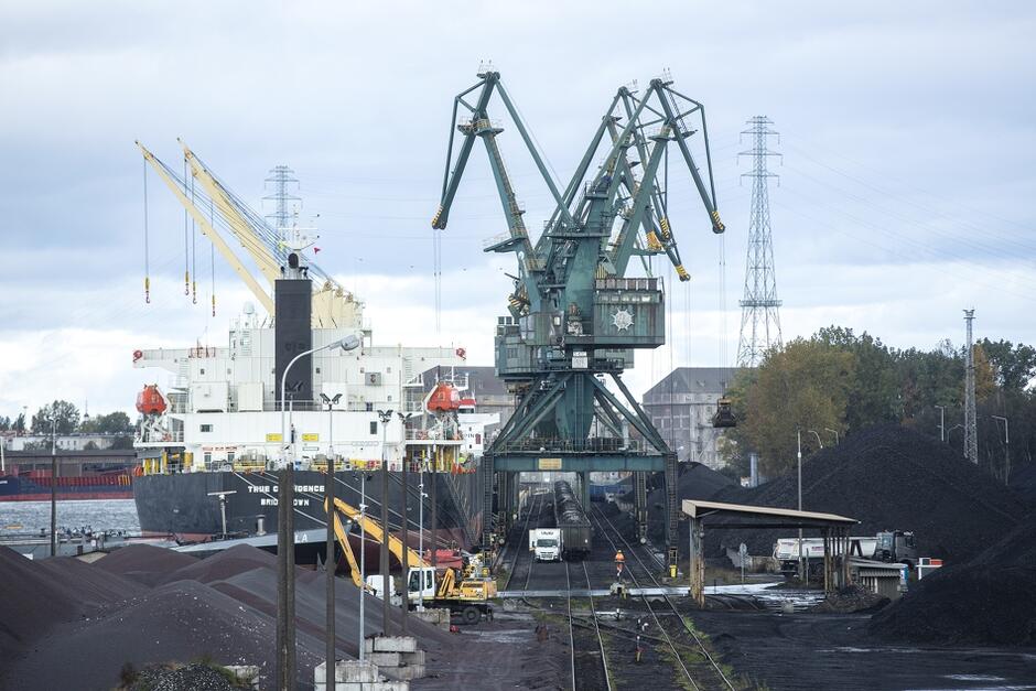 na zdjęciu teren portu morskiego, widać ogromny dźwig portowy, pod nim wagony kolejowe wypełnione węglem, wokół wysokie hałdy węgla, w tle statki